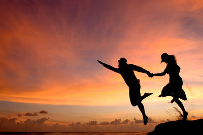 Silhouette couple holding hands while jumping in sea against sky during sunset