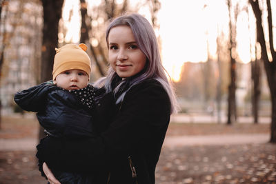 Happy young mother carrying baby son standing in park in autumn