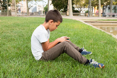 Side view of boy using phone while sitting on grassy land