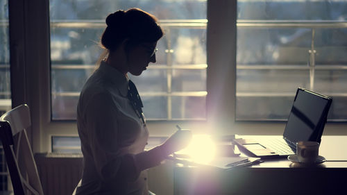 Dark silhouette of young focused attentive businesswoman wearing glasses, writes something in tablet