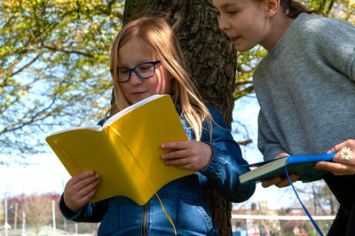 Two young smiling school age girls meet in the park and read from yellow textbook.