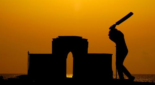 Silhouette man playing with bat against sky during sunset