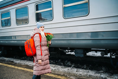 Girl on the platform of the station with a red backpack and a bouquet of tulip flowers