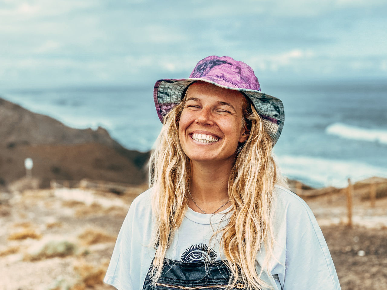 one person, smiling, portrait, happiness, land, adult, women, spring, long hair, beach, hat, emotion, nature, blond hair, clothing, teeth, leisure activity, hairstyle, smile, water, sea, trip, vacation, holiday, cheerful, young adult, blue, front view, sky, casual clothing, female, day, portrait photography, looking at camera, fashion accessory, enjoyment, focus on foreground, travel, sand, headshot, photo shoot, lifestyles, landscape, standing, outdoors, travel destinations, carefree, fun, waist up, relaxation, environment, positive emotion, summer, looking, person, sun hat, fashion, beauty in nature, rural scene, sunlight