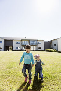 Full length portrait of siblings holding hands while walking at lawn outside house against clear sky