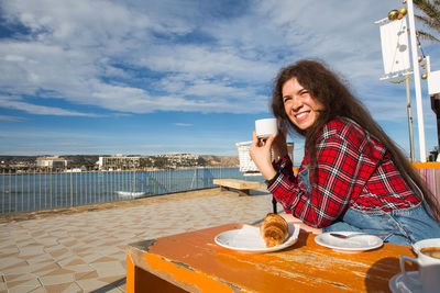 Woman holding coffee while sitting on table against sky