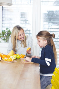Happy mother with orange looking at daughter cutting fruit on table at home