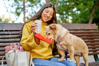 Positive young female owner giving water to cute dog with special device while sitting on wooden bench on summer day in park