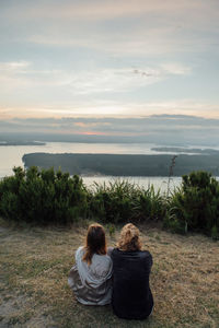 Rear view of friends sitting on hill by sea against sky