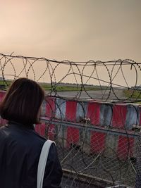 Rear view of woman standing by fence against sky during sunset