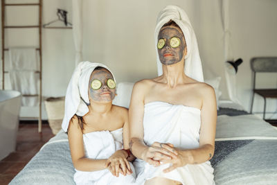 Mother and daughter in towel with facial mask and slice of cucumber on eyes sitting at home