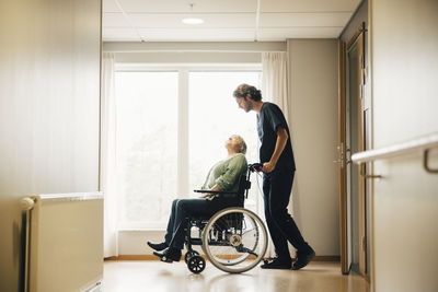 Full length side view of male nurse pushing disabled senior woman on wheelchair in alley at retirement home