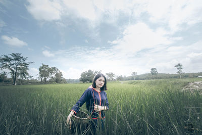 Young woman carrying basket while standing amidst crops on field against sky