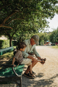Grandfather helping grandson wearing sandal while sitting on bench at park