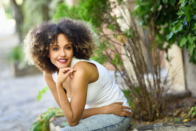 Portrait of smiling young woman sitting on retaining wall by plants