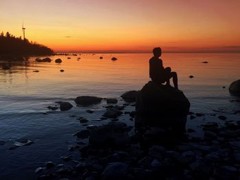 Silhouette person sitting on rock at beach during sunset