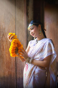 Smiling woman holding floral garland standing against wall