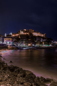 Illuminated city by sea against sky at night