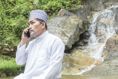 Thoughtful man talking on phone while sitting against waterfall
