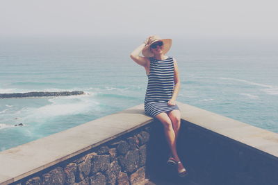 Portrait of woman sitting on retaining wall by sea against sky