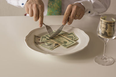 Midsection of man cutting paper currencies in plate with fork and table knife