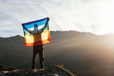 Back view of hiker standing with rainbow lgbt flag with inscription peace and enjoying freedom with outstretched arms in mountains