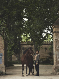 Full length of adult black female in elegant outfit standing with brown horse near green trees and castle fence in daytime in yard