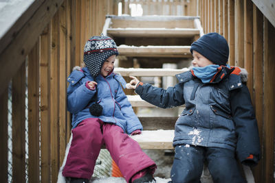 Happy siblings playing with snow while sitting on steps during winter
