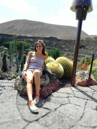 Portrait of smiling young woman sitting on rock against sky during sunny day