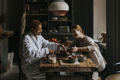 Smiling non-binary person sharing smart phone with woman having breakfast at home