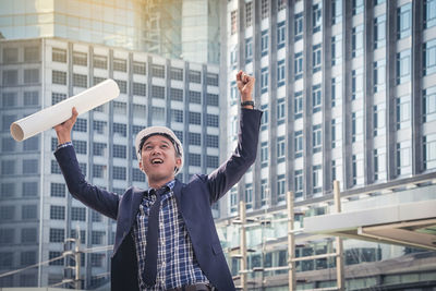 Happy businessman with arms raised standing against buildings