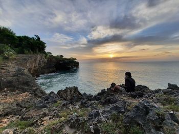 People sitting on rock by sea against sky during sunset