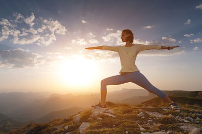 Young woman in warrior yoga pose standing on mountain rock under beautiful cloudy sky