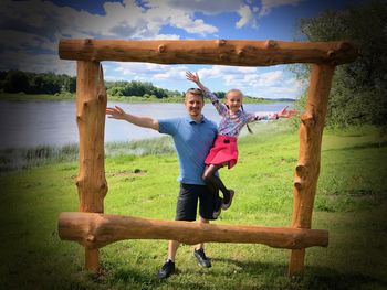 Father and daughter standing in front of wooden frame on field by lake