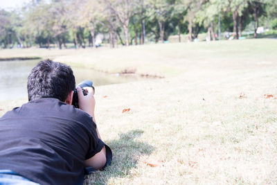 Rear view of man photographing trees while lying on grass at park