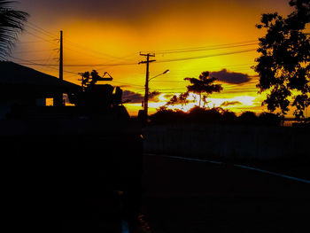 Silhouette trees by road against orange sky