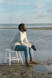 Relaxed young man with eyes closed sitting on white stool at beach against sky