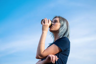 Blue haired woman drinks take away coffee against blue sky.