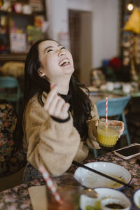 Young woman laughing while holding drink sitting at restaurant