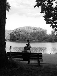 Rear view of woman sitting on bench by lake against sky