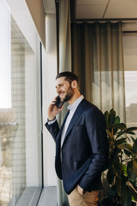 Smiling businessman talking on mobile phone while standing near window in coworking office