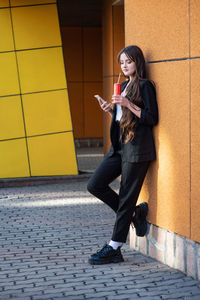 Teenager girl in black suit stands near orange wall, drinks soda from can, uses phone youth culture