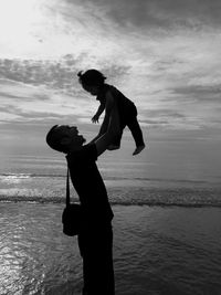 Side view of silhouette man carrying baby girl at beach against sky