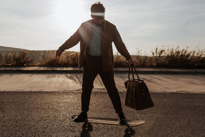 Full body serious young bearded male skater in trendy clothes with leather bag riding skateboard along asphalt road