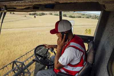 Side view of male combine operator talking on smartphone while harvesting wheat grains in professional industrial machine in agricultural field