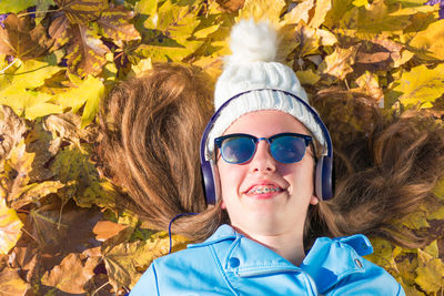 Teenager girl wearing sunglasses and cap is lying down on the floor covered by yellow leaves while enjoying listening to music by headphones. the brunette girl is happy and smiling. horizonta