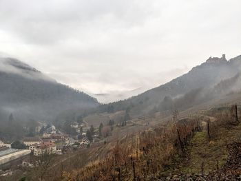 A misty view of a town in a valley with a castle on a mountain in ribeauville, france. 