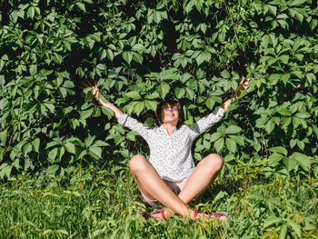 Wide smiling woman sits on grass lawn near wall all over covered with thick ivy. summer vibes.