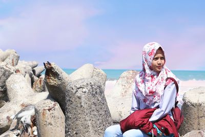 Woman wearing hijab sitting on rock against sea