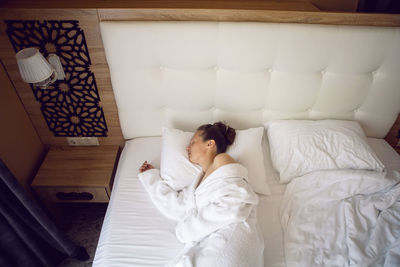 Woman in a white bathrobe is sitting on the edge of the bed and drinking tea from mug, large window 